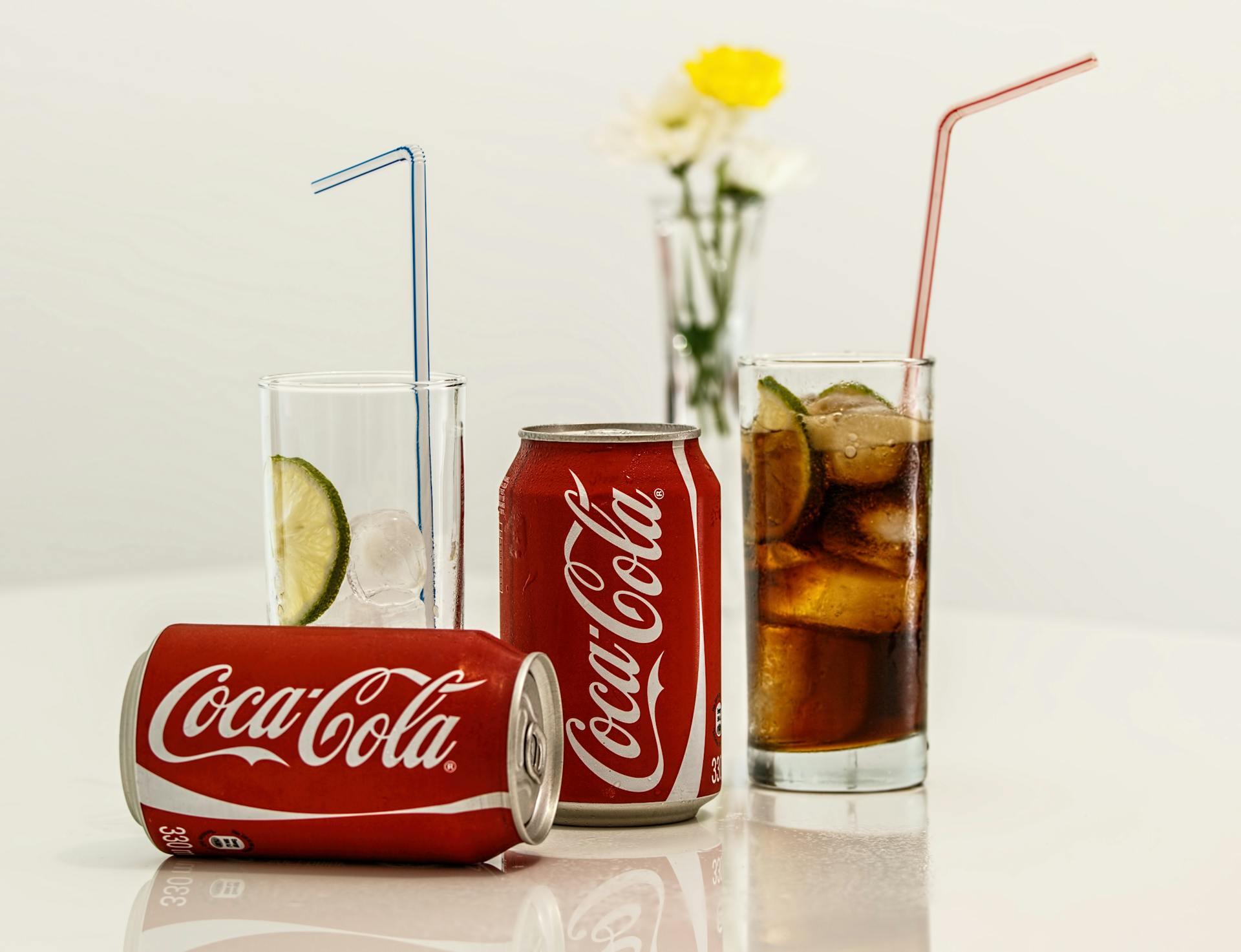 Two cans of Coca-Cola, one on its side and one upright, on a table next to one empty and one full glass with straws, a vase of yellow flowers in the background