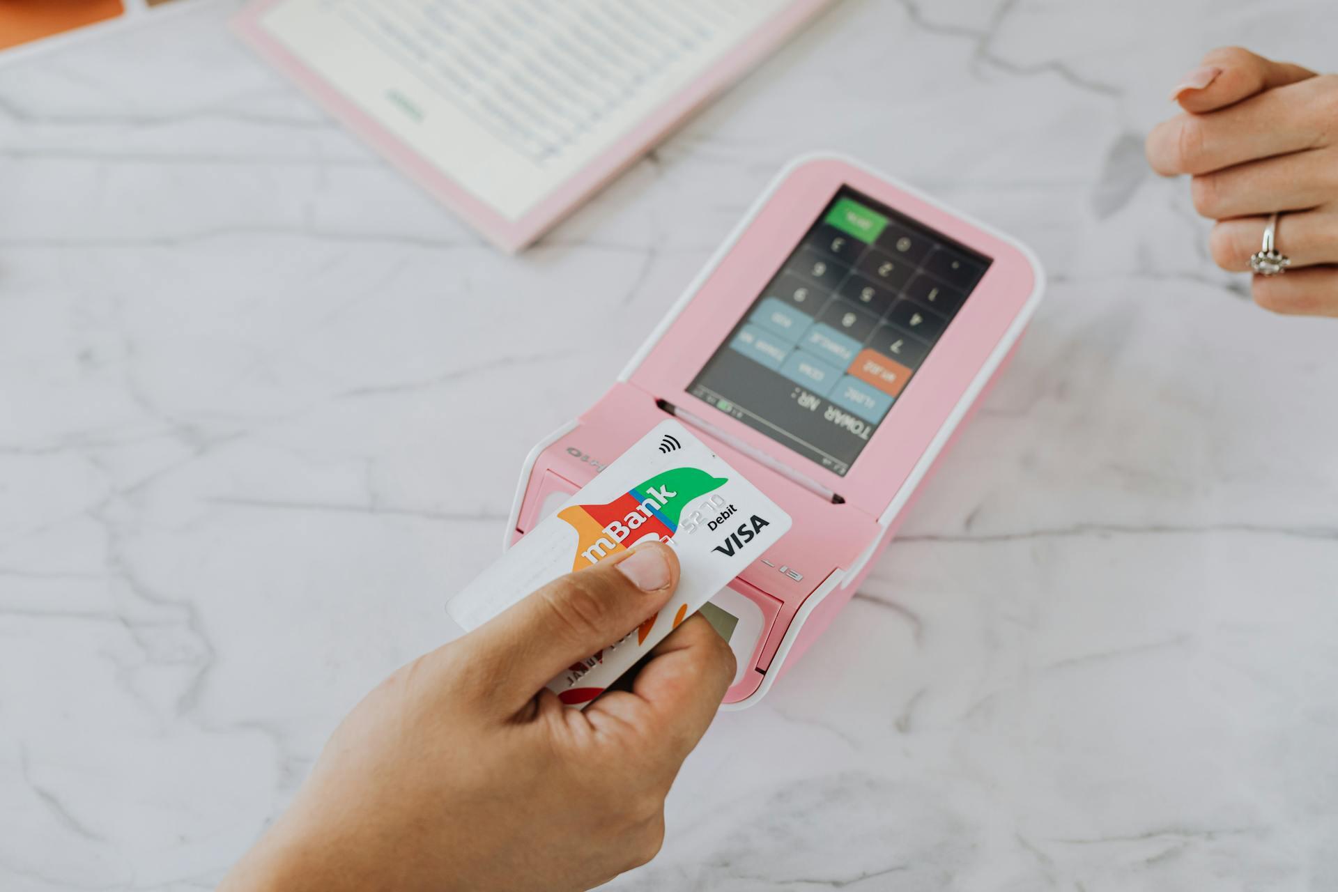 A hand holding an mBank Visa debit card above a pink payment terminal on a marble surface, with another hand and pink notebook nearby