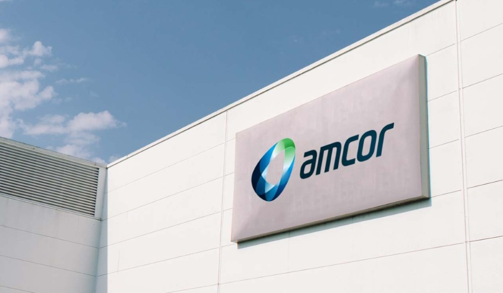 A grey sign reading "amcor" on the inner corner of two white walls, blue sky above