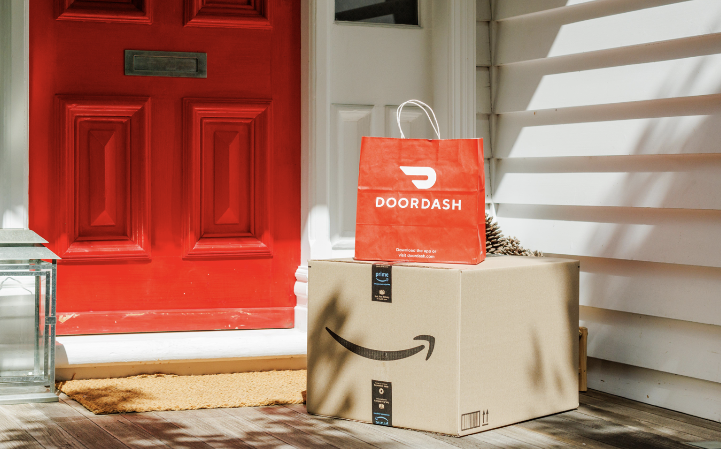 An orange shopping bag reading "DOORDASH" on top of a brown cardboard box with Amazon's smile logo, on a front doorstep near a door painted orange