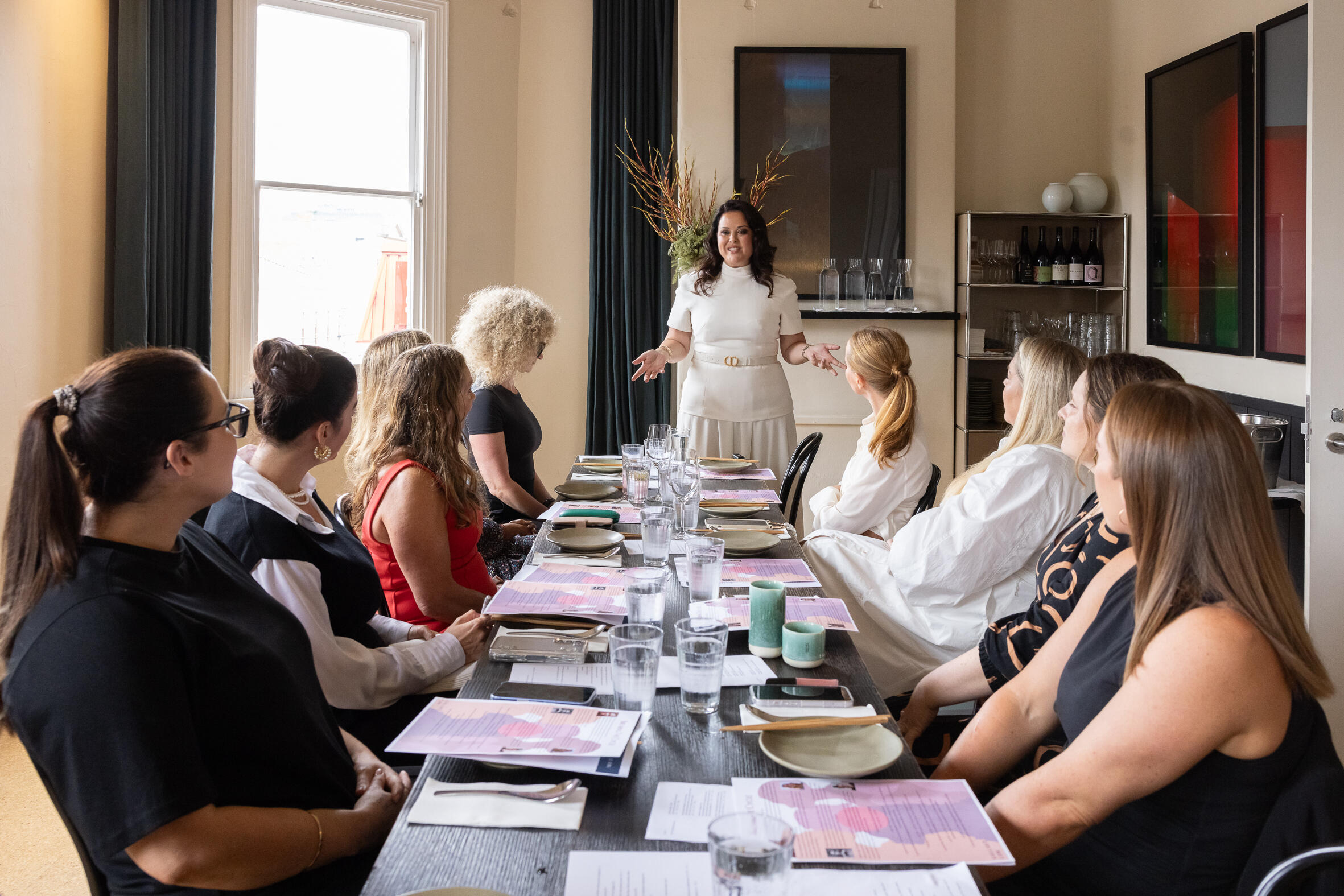 A woman in a belted white dress speaking while standing at the end of a long dining table seating several other women