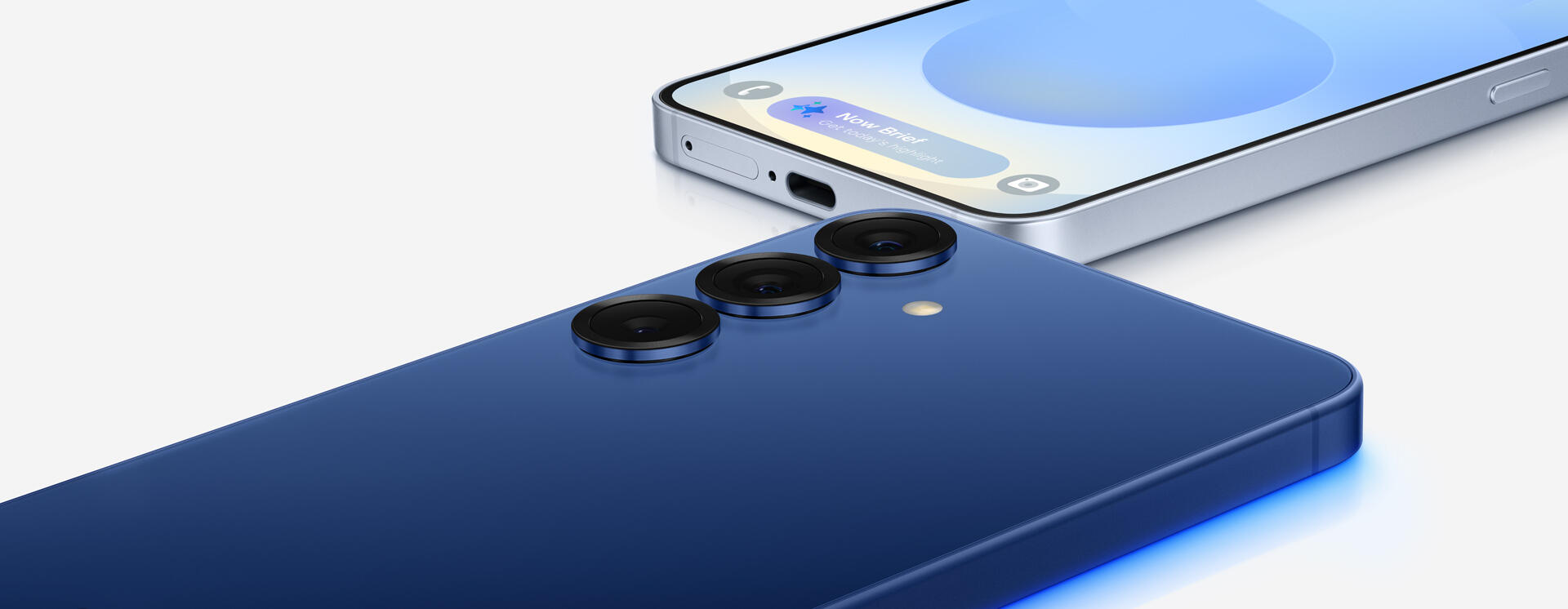 A dark blue smartphone with three rear cameras lying on its screen next to another smartphone lying on its back