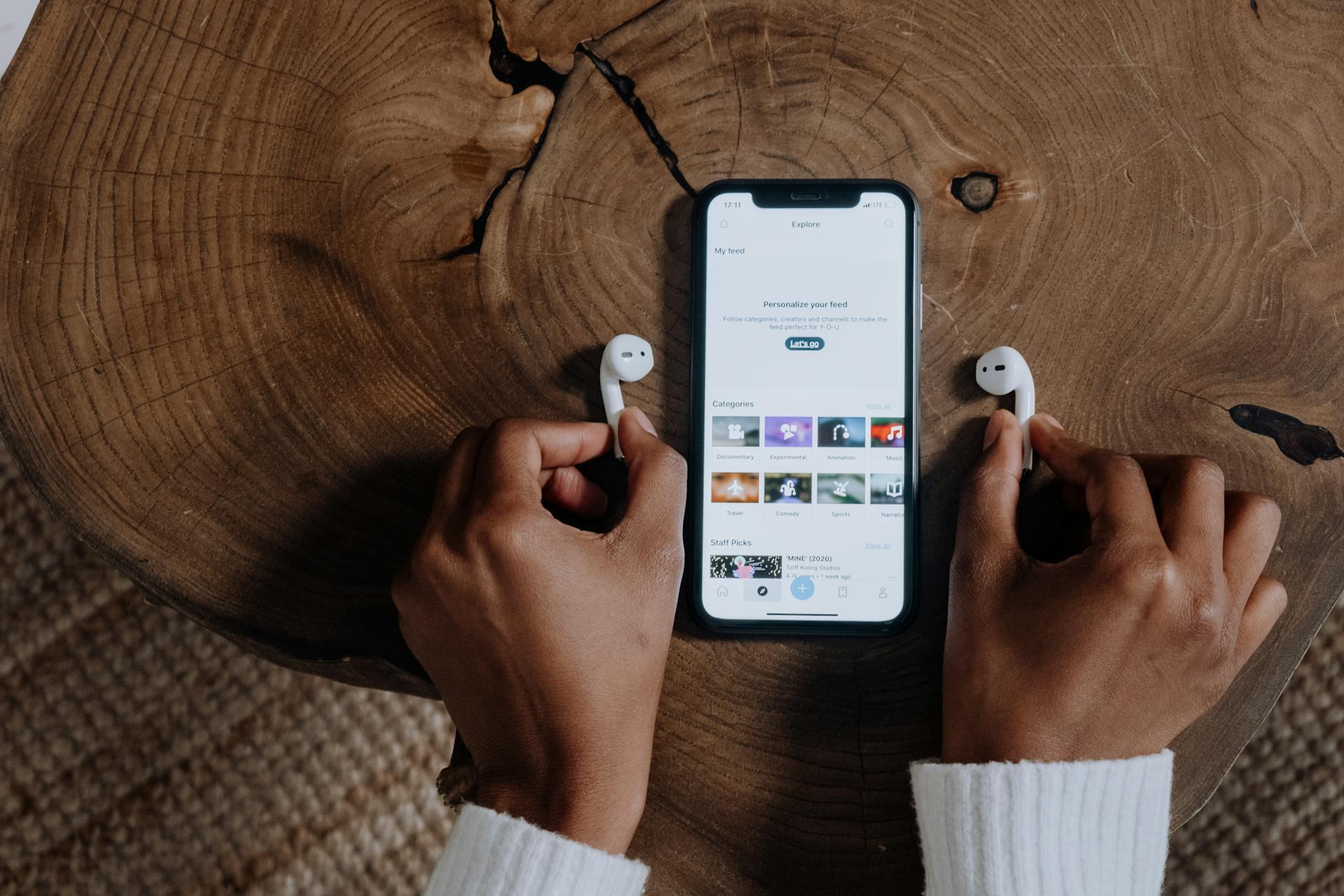 Hands holding white earbuds next to a phone showing a streaming service, on a wooden table