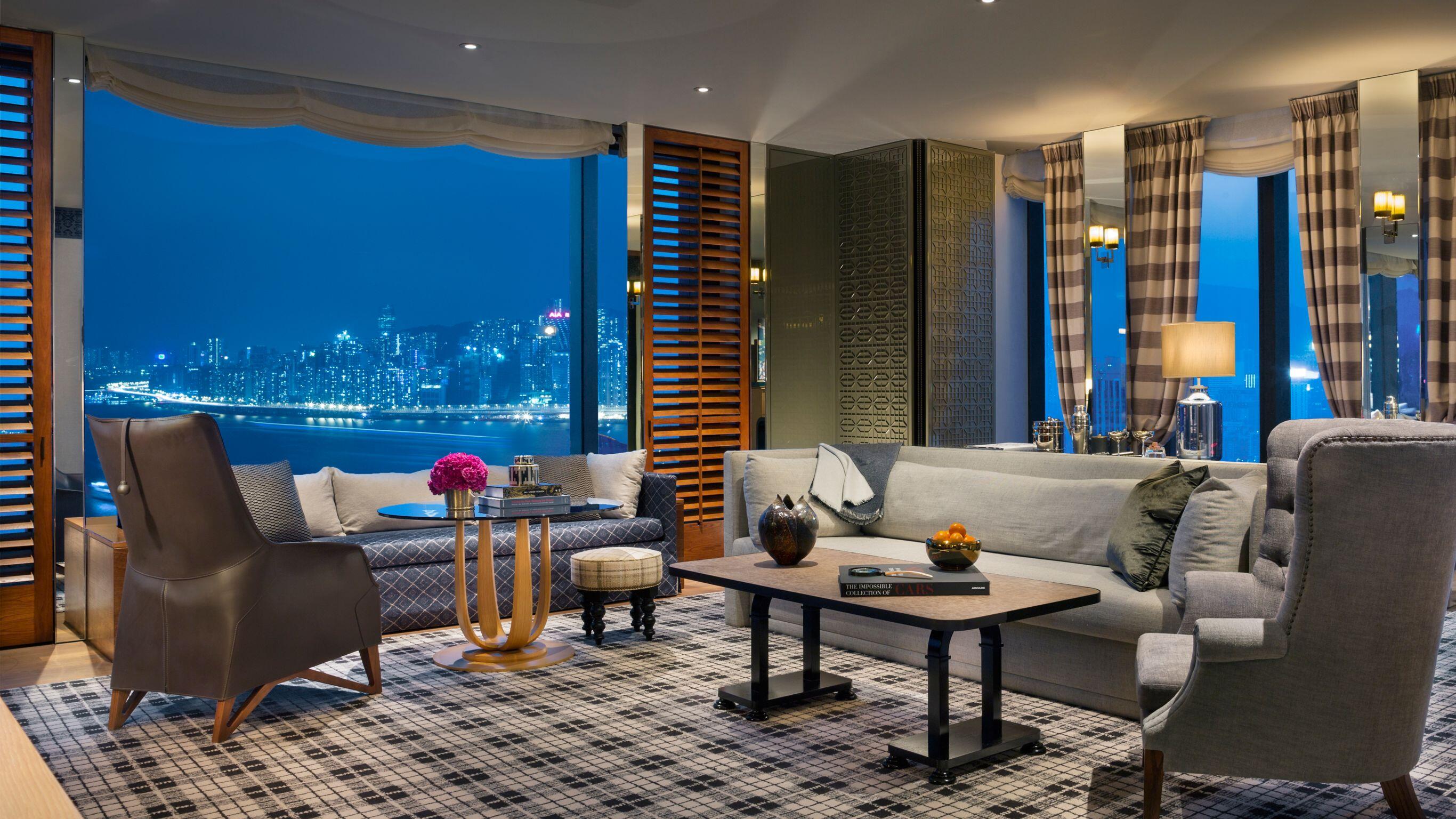 A hotel room living area with couches, chairs, and tables in front of windows to the Hong Kong skyline at night