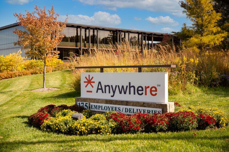 A white sign saying "Anywhere" and "175, employees/deliveries" in front of orange-leafed trees and an office building