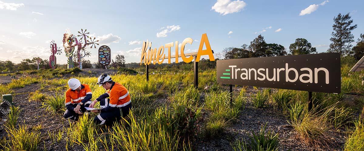 Two people in orange hi-vis jackets crouching in the grass next to signs that say "Transurban" and "Kinetica"